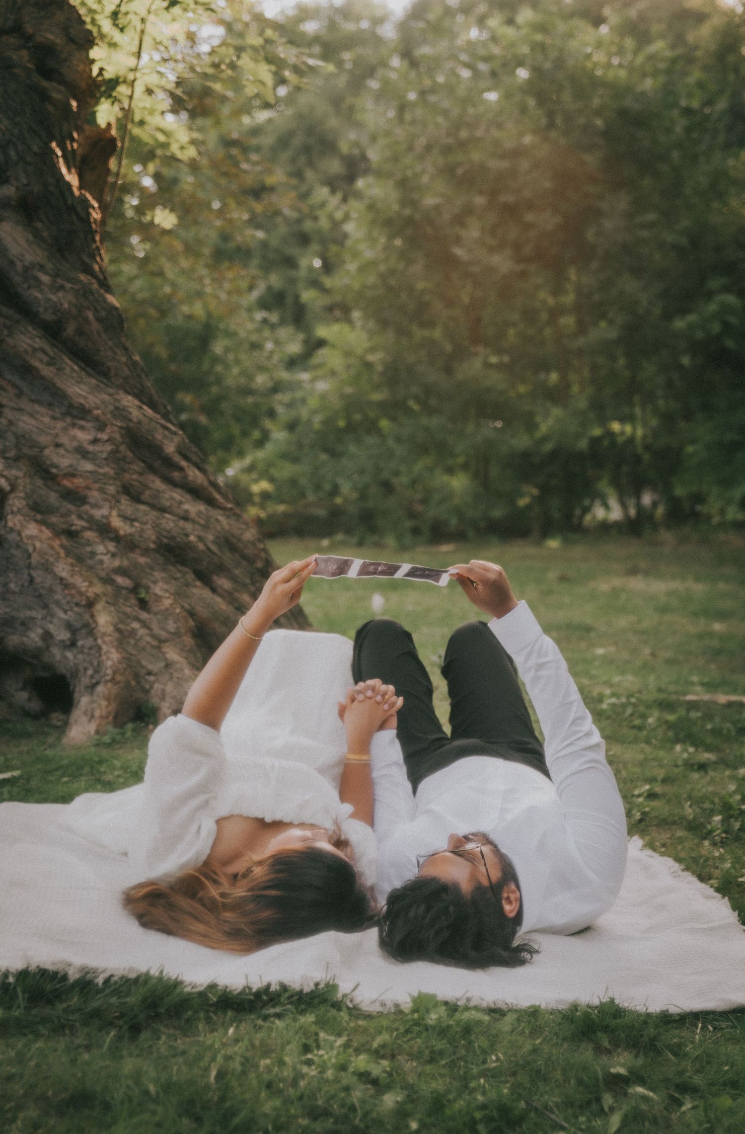 A couple lies on a white blanket in a park looking at something together.