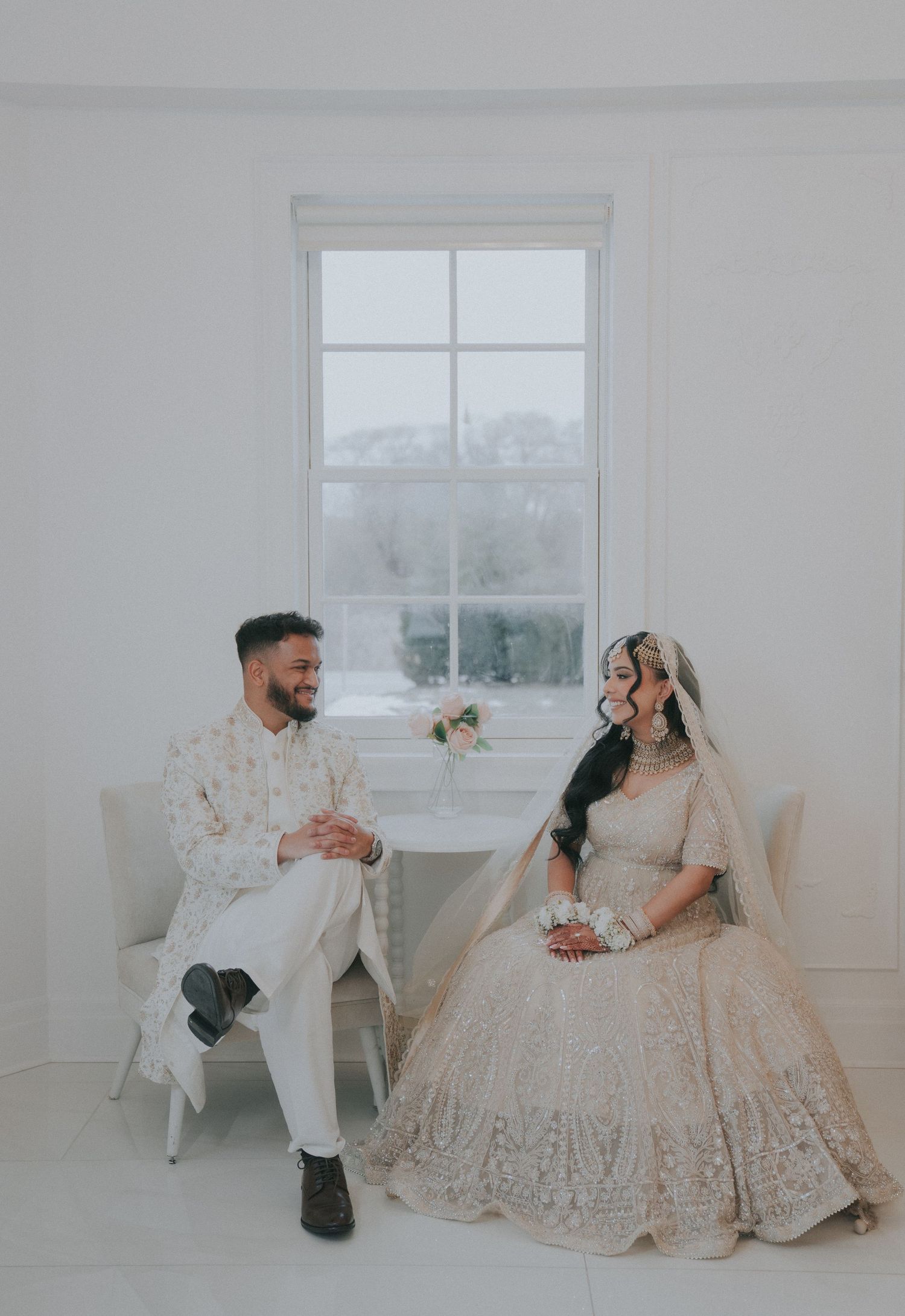 Elegant wedding couple poses in a minimalist white room with window light creating romantic atmosphere.