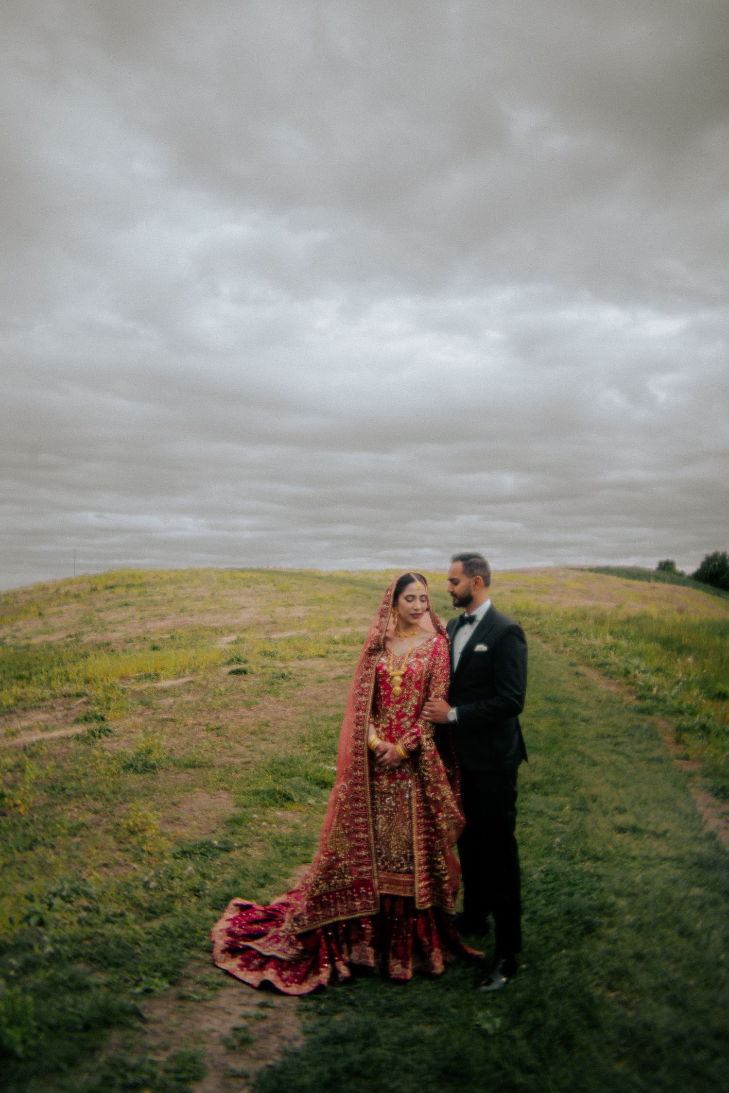 Couple in formal attire standing on a grassy hillside under moody skies.