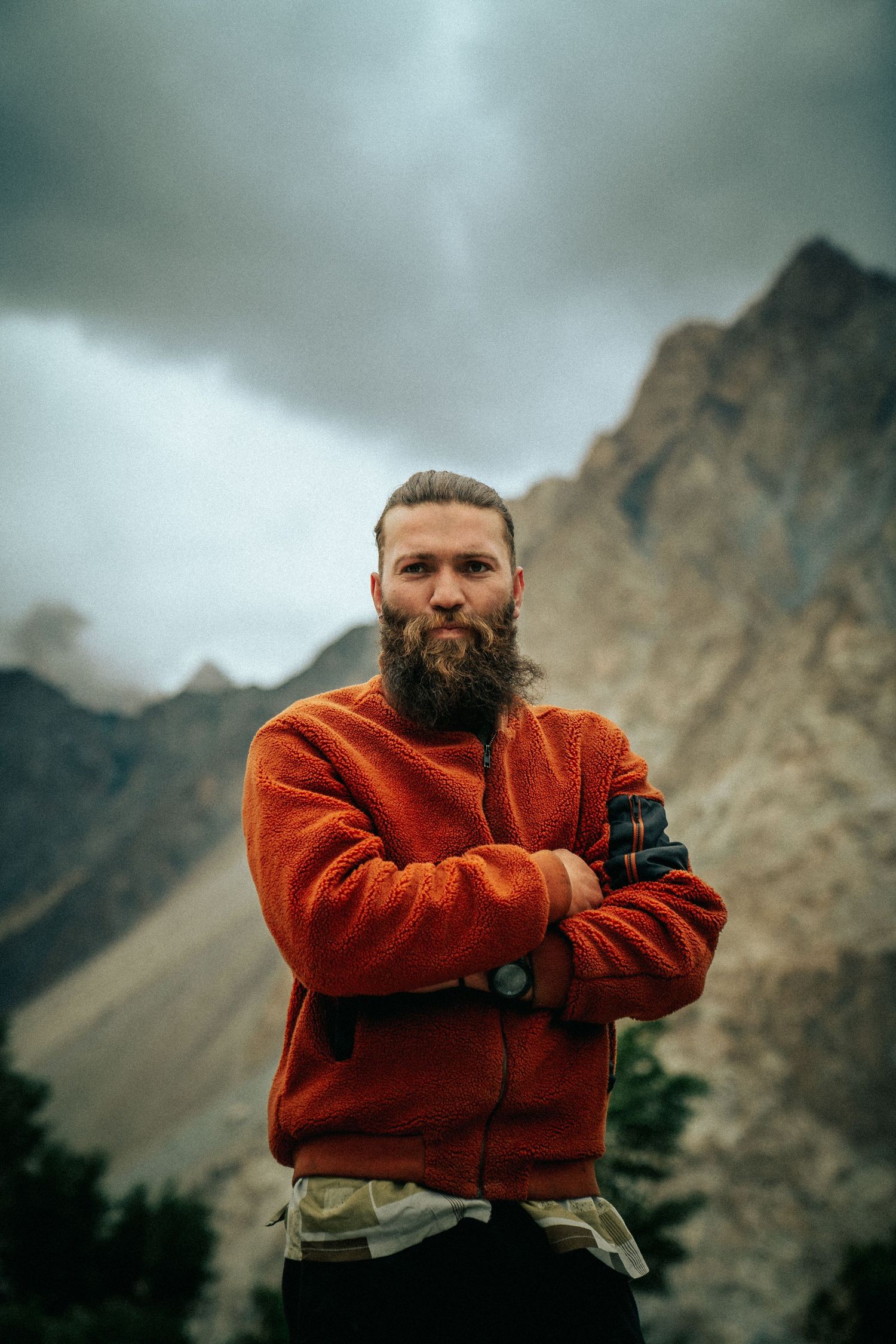 Person in orange fleece jacket poses thoughtfully against dramatic mountain landscape.