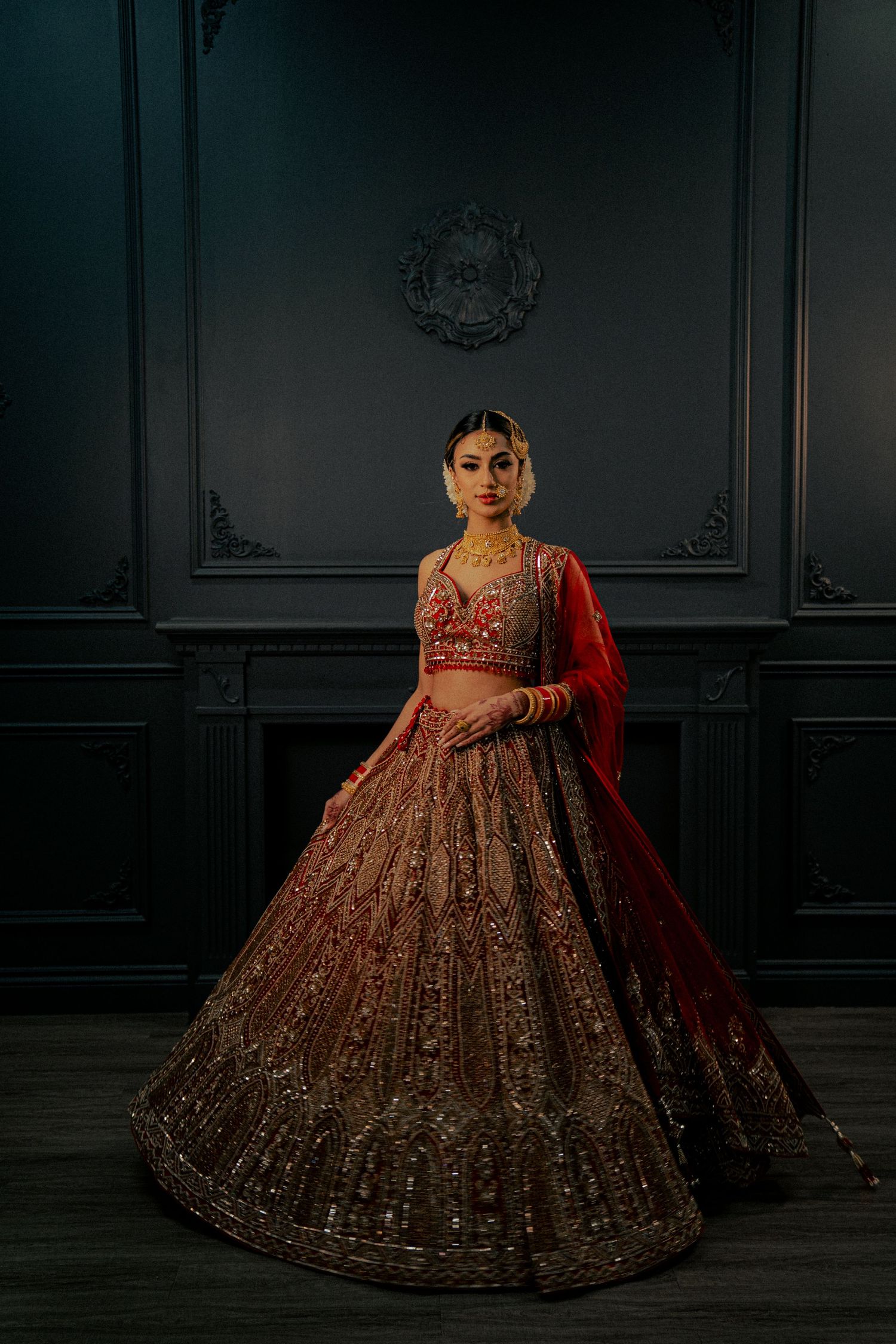 A dramatic bridal portrait in a dark room featuring an ornate red and gold lehenga.