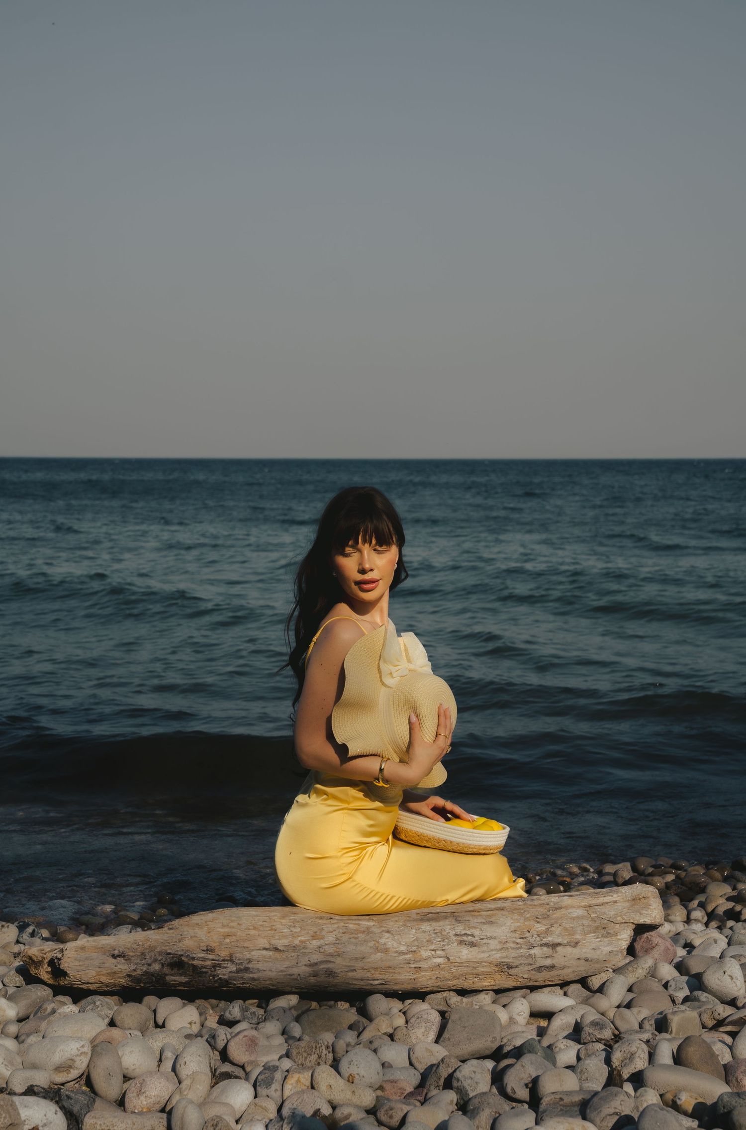 Person in yellow dress sitting on rocky beach beside ocean at dusk.