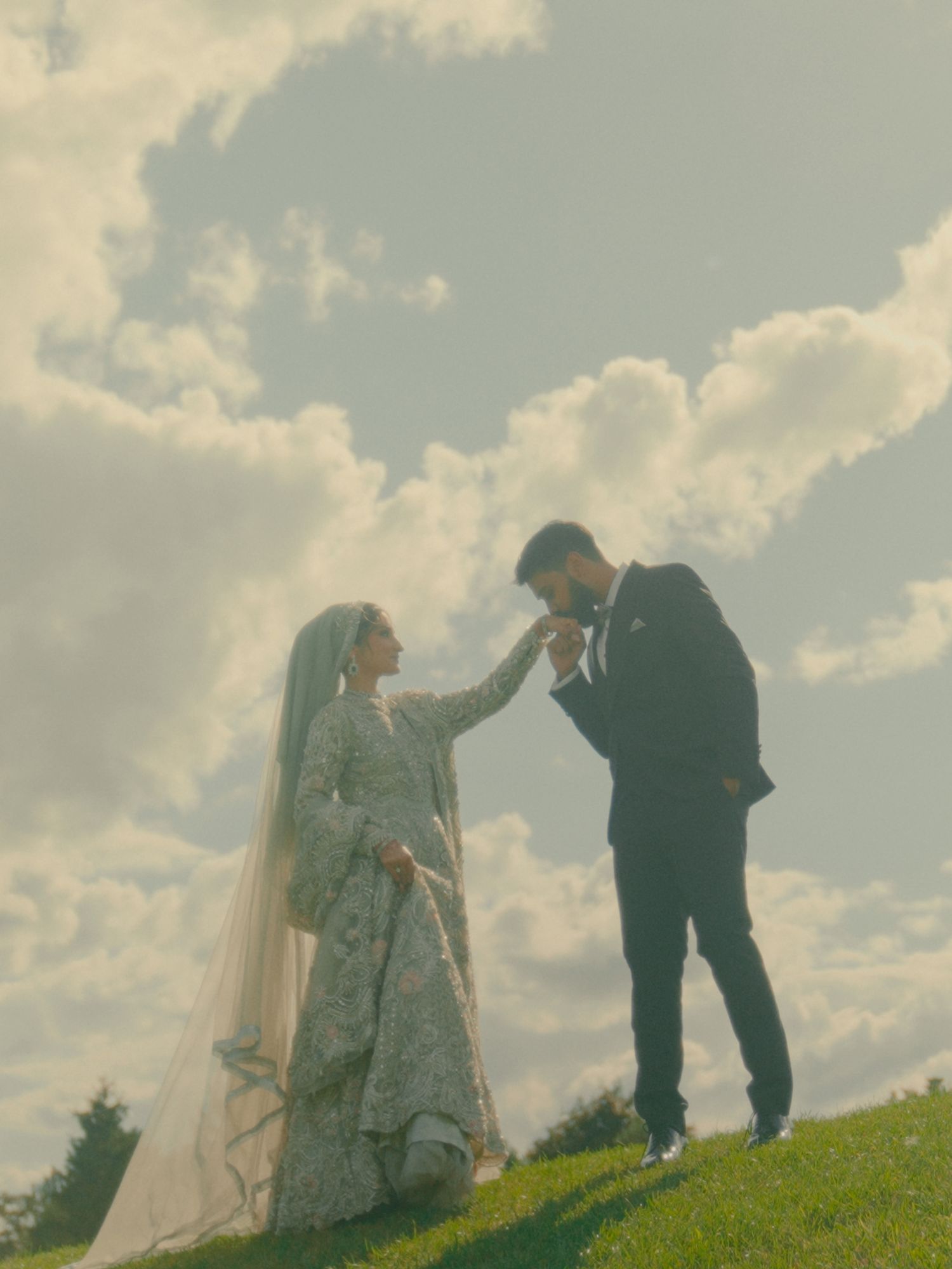 Couple in formal wear standing on a grassy hillside under soft clouds.