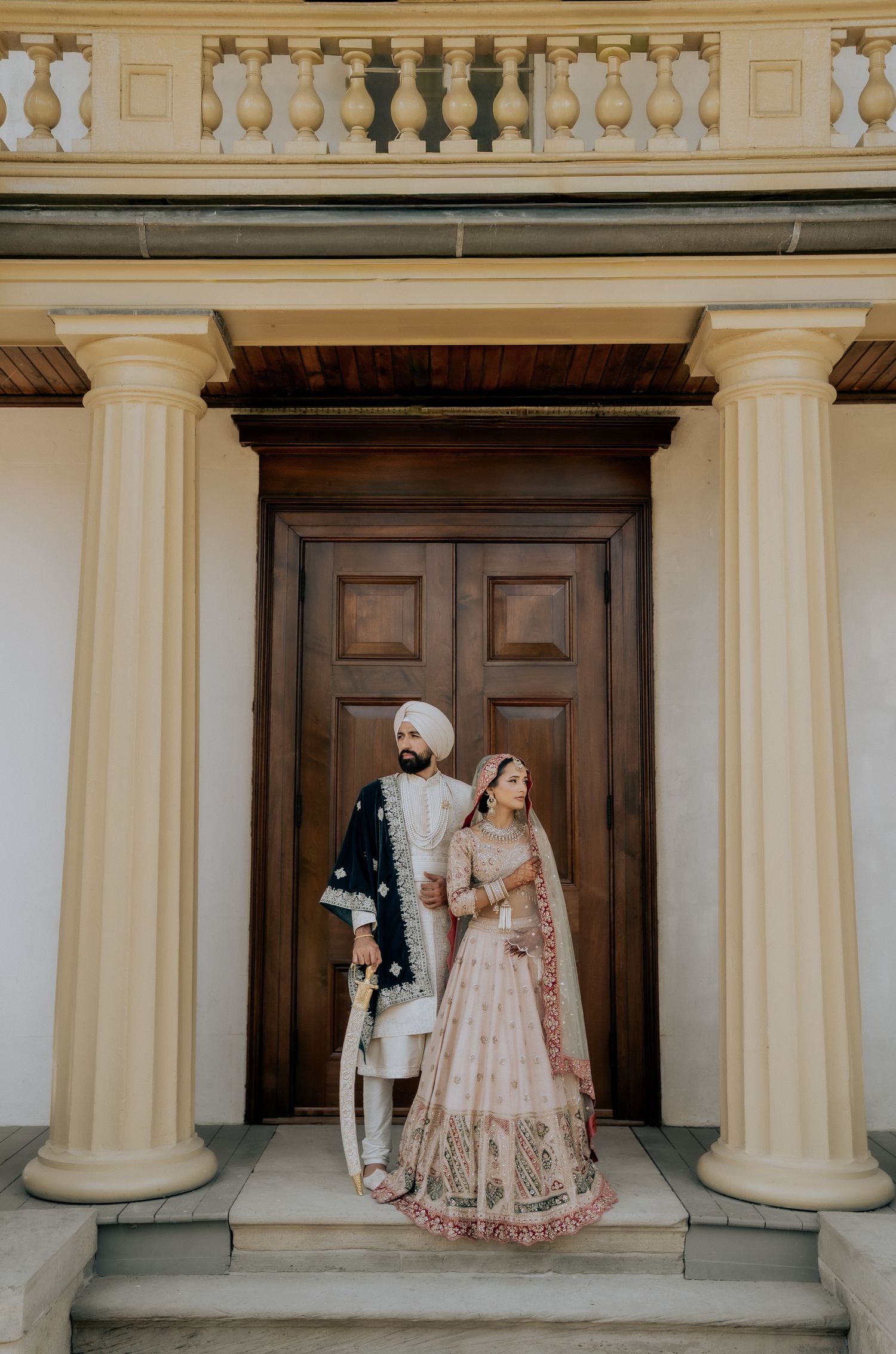 Elegant couple poses between ornate white columns in traditional South Asian wedding attire.