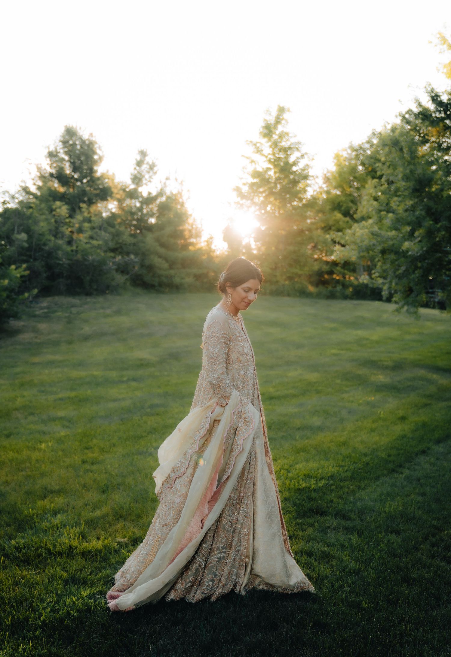 Ethereal bridal portrait in flowing cream dress against sunset and trees.