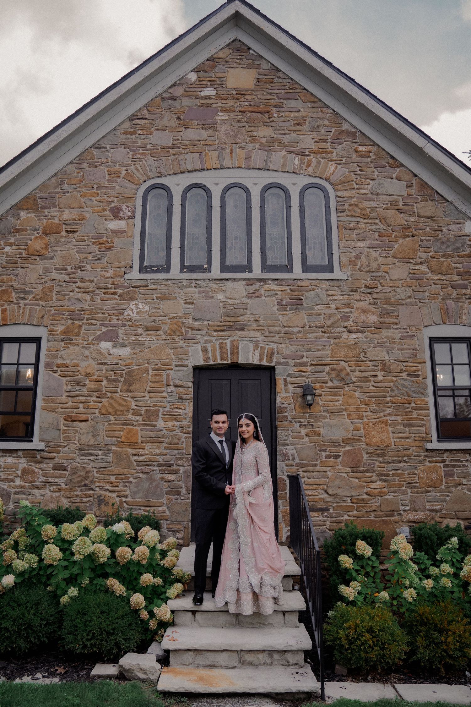 Newlyweds pose on stone church steps surrounded by hydrangea bushes.