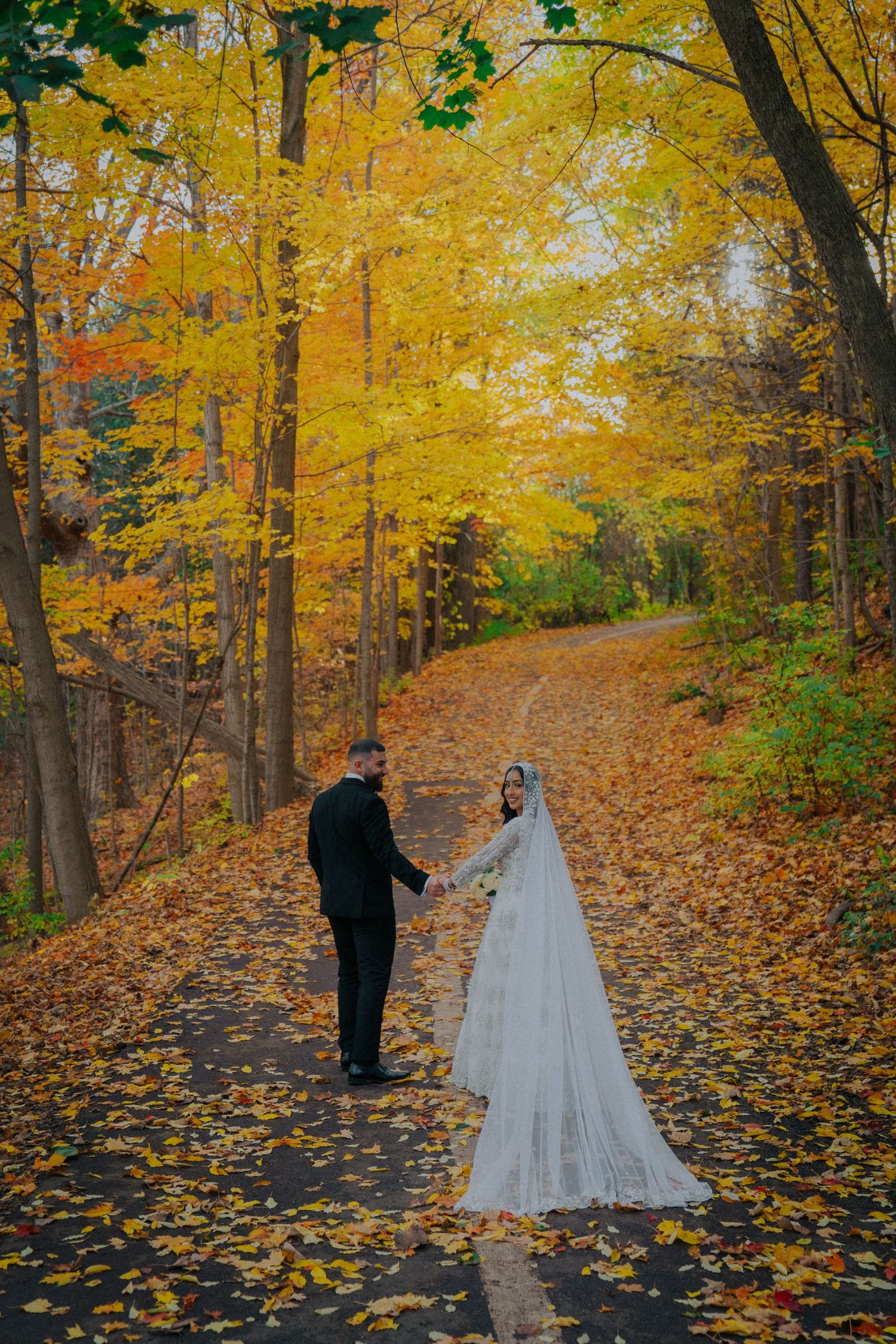 Wedding couple walks through vibrant autumn forest path covered in golden fallen leaves.