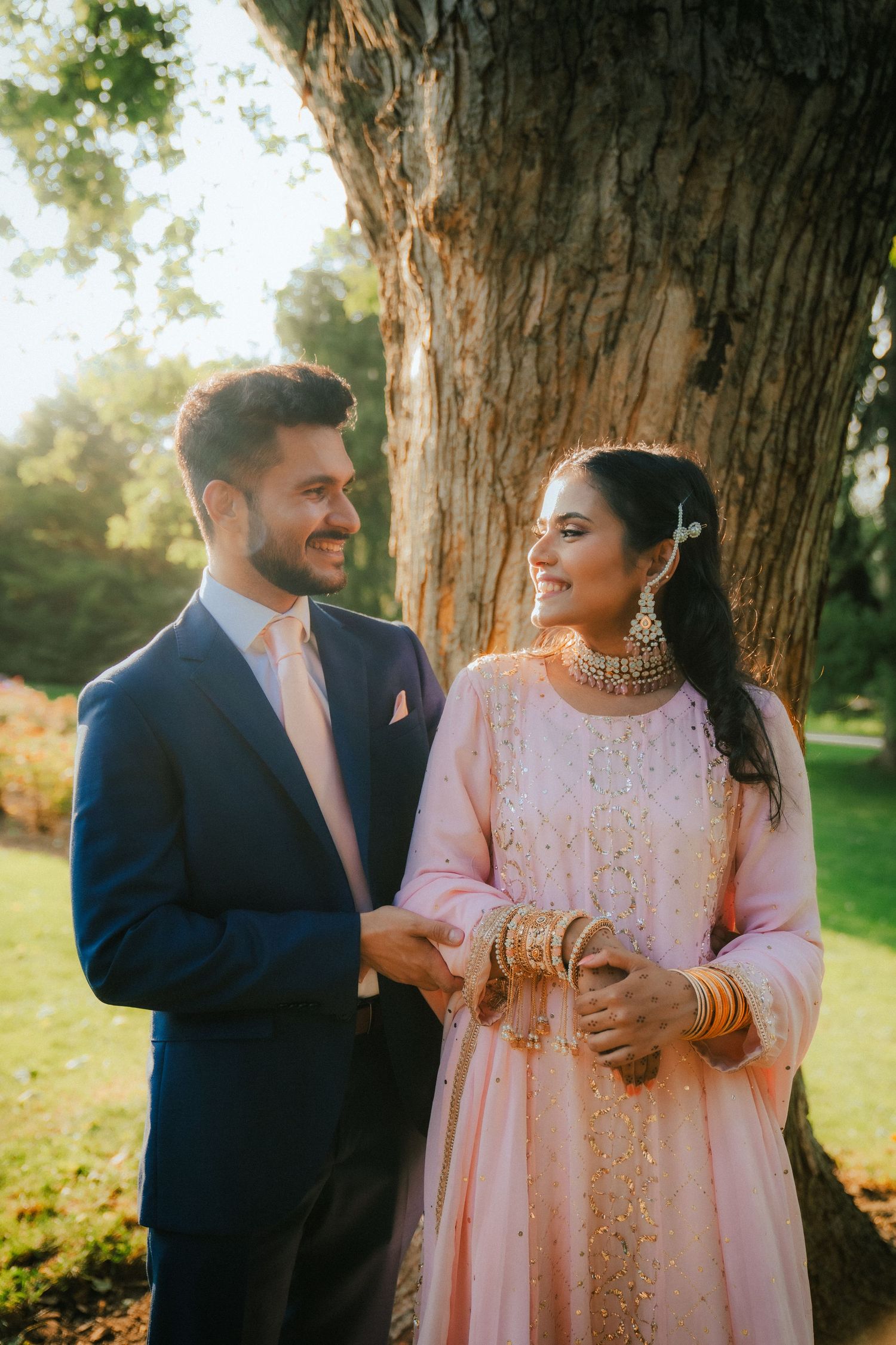Couple in traditional South Asian wedding attire posing under a large tree.