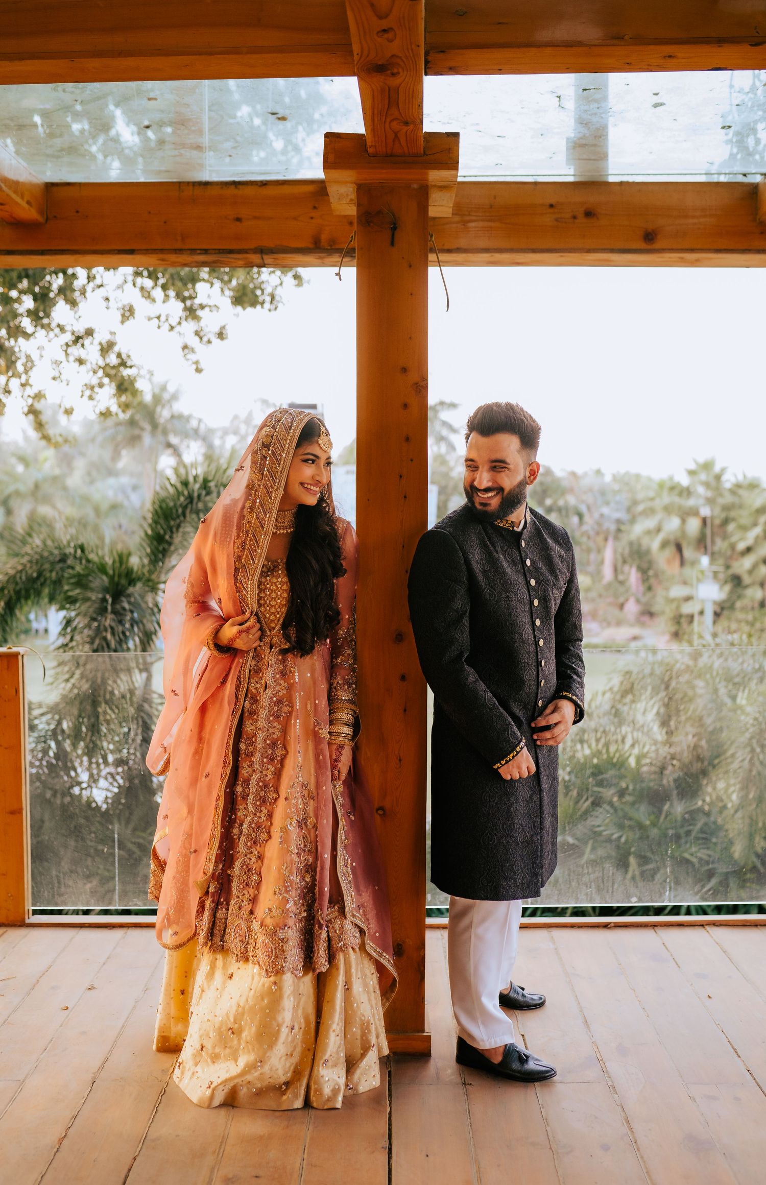 A couple in traditional South Asian wedding attire stands together on a wooden deck with natural lighting and rustic beams above.