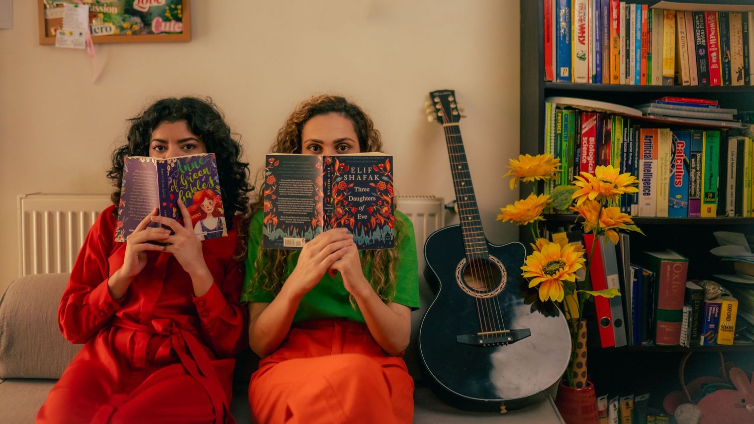 Two readers hide behind books in a cozy room with a guitar and sunflowers against a bookshelf backdrop.