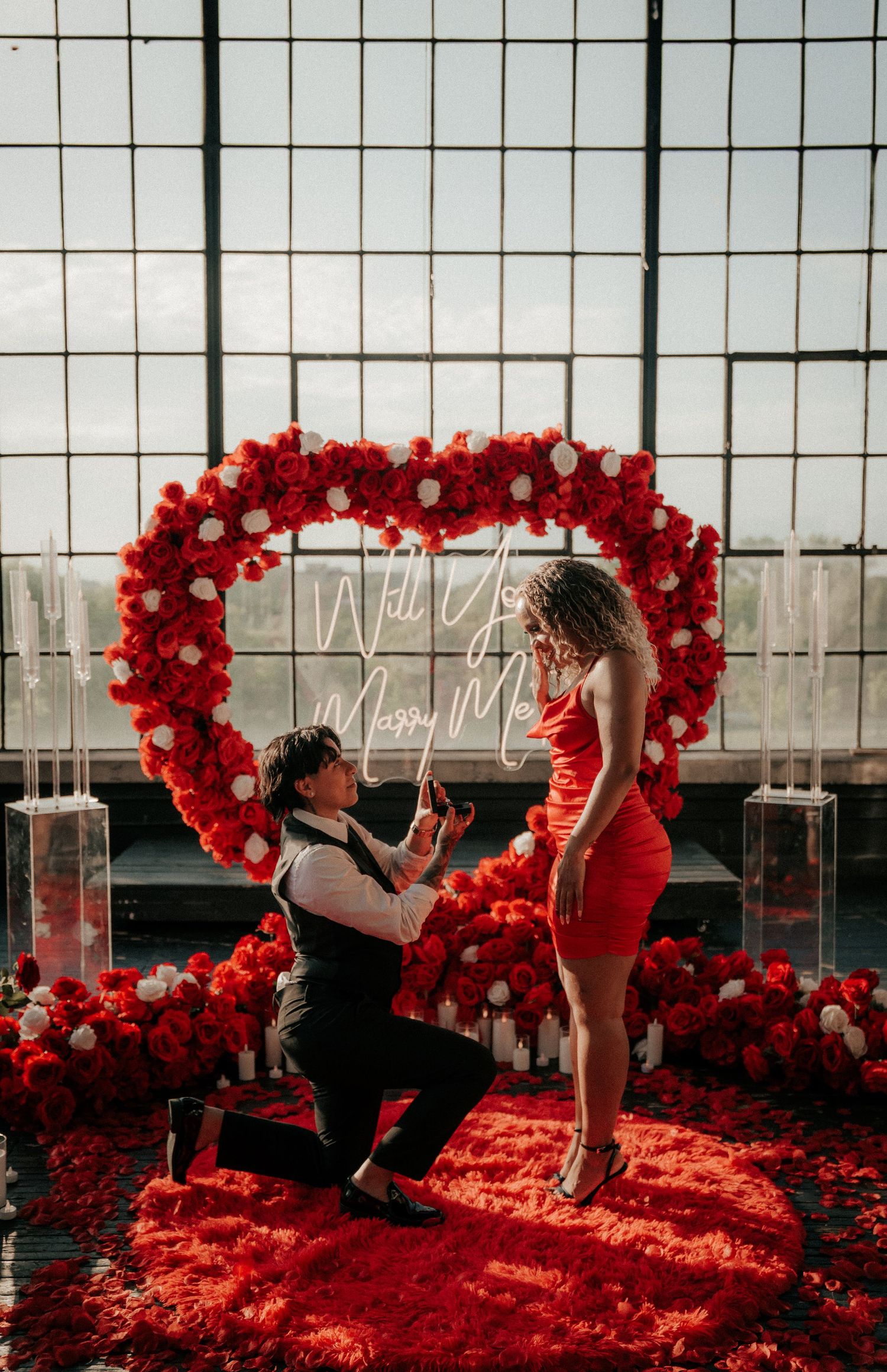 A romantic proposal setup with red balloon heart arches and rose petals in a modern glass-walled space.