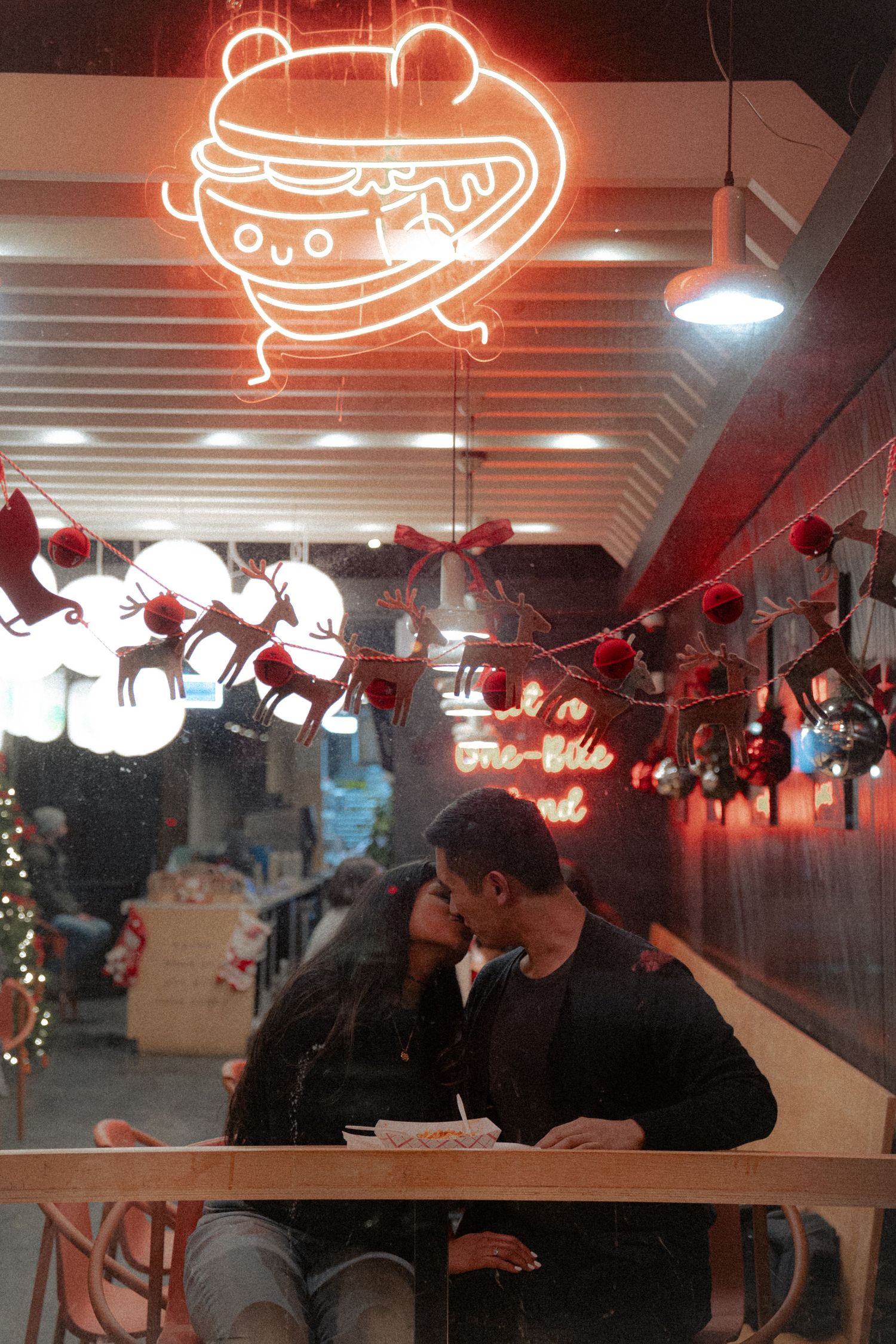 A romantic moment under a neon burger sign in a dimly lit restaurant setting.