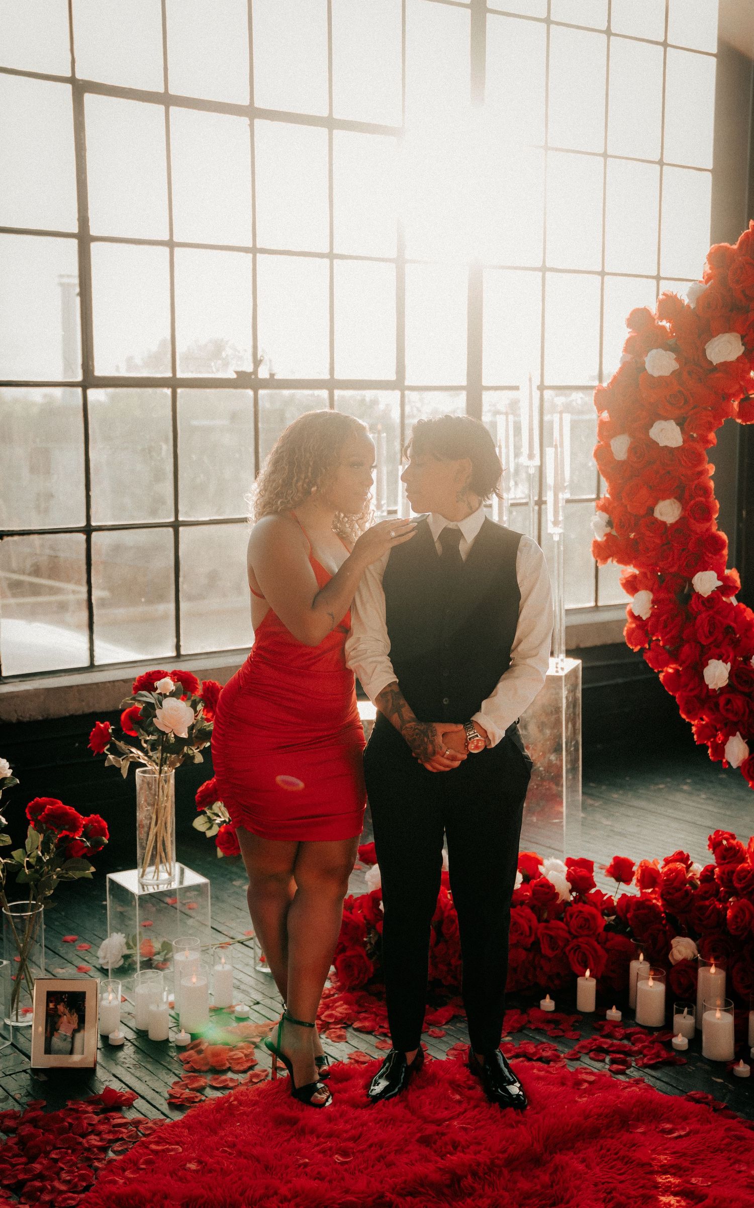 A couple stands amid red rose petals and balloon decorations in a sunlit industrial space.