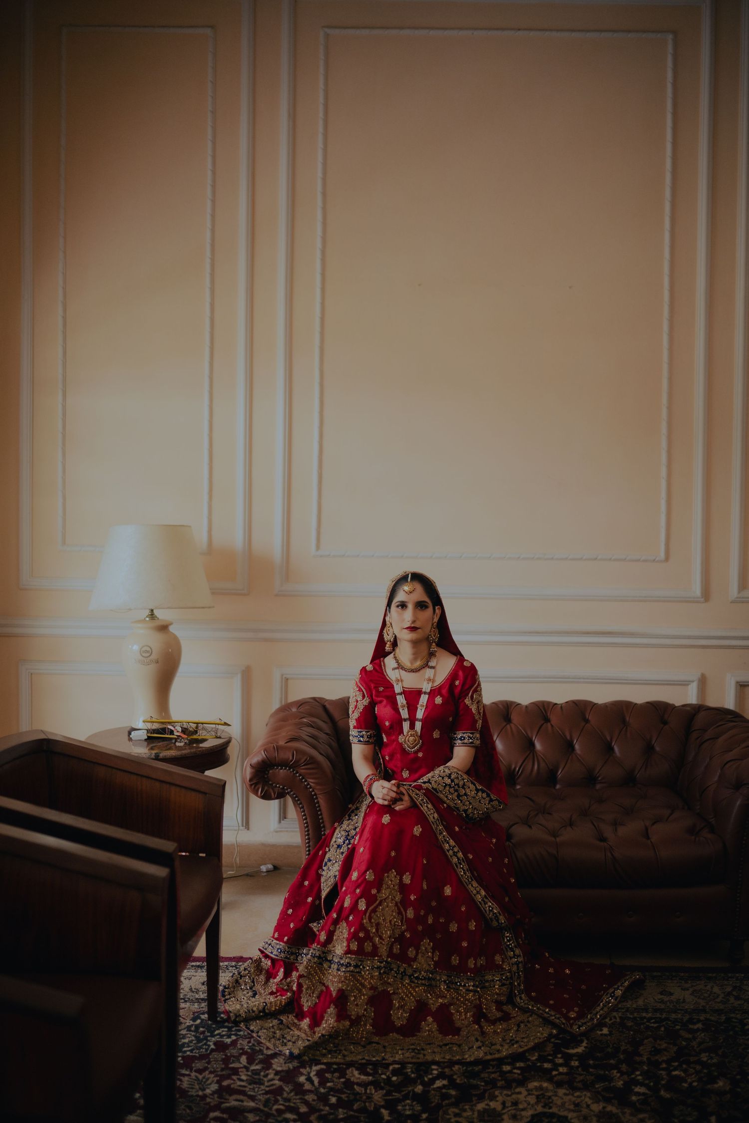 Traditional South Asian bridal attire featuring red and gold lehenga photographed in elegant interior setting.