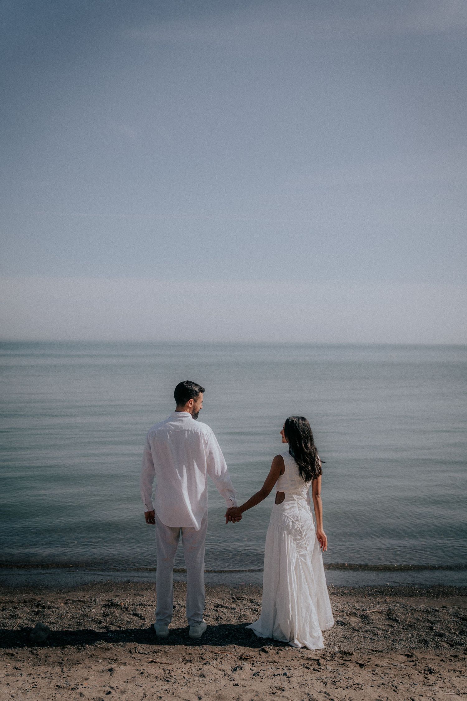 A couple in white attire walks along a serene beach shoreline.