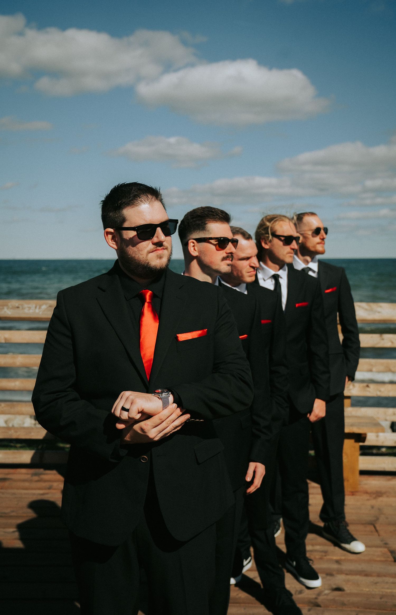 Groomsmen in black suits and red ties pose by oceanside deck.