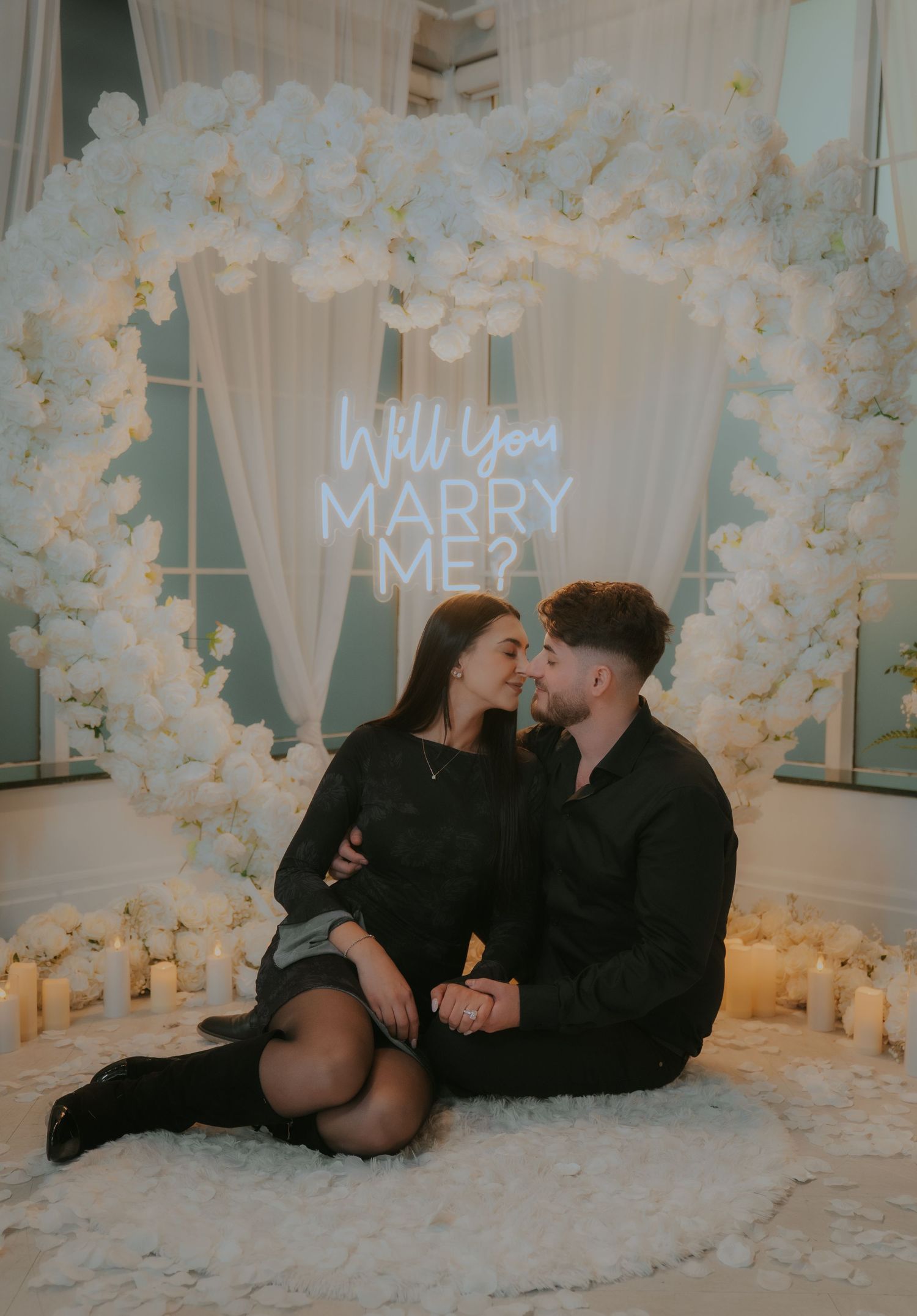 A romantic proposal scene with white floral decorations and illuminated text in the background.