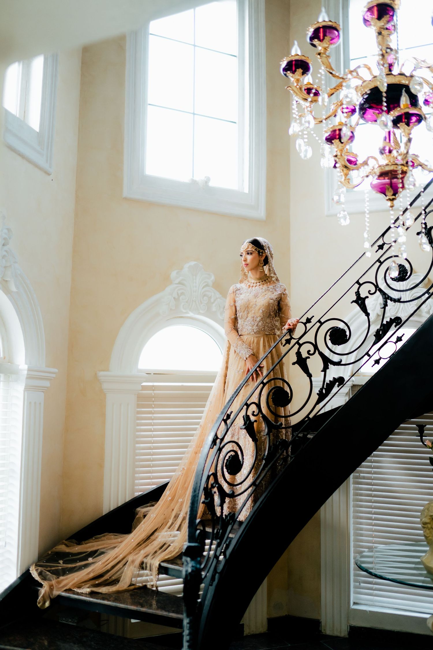 Ornate bridal portrait on grand staircase with elegant chandelier and white interior.