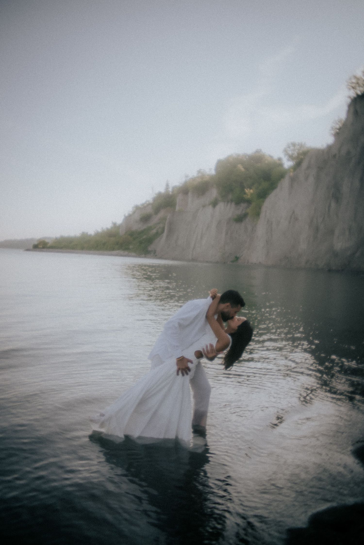 A romantic couple in wedding attire poses dramatically near cliffs by the water.