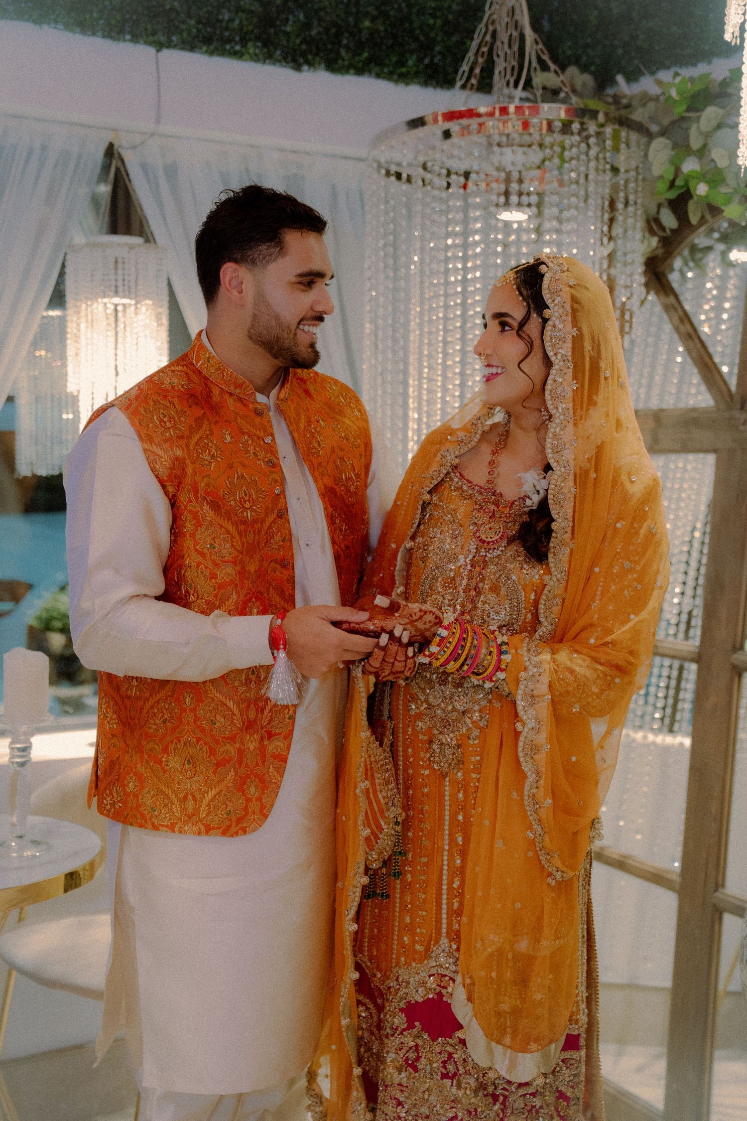 A couple in traditional orange and gold South Asian wedding outfits sharing tender moments.