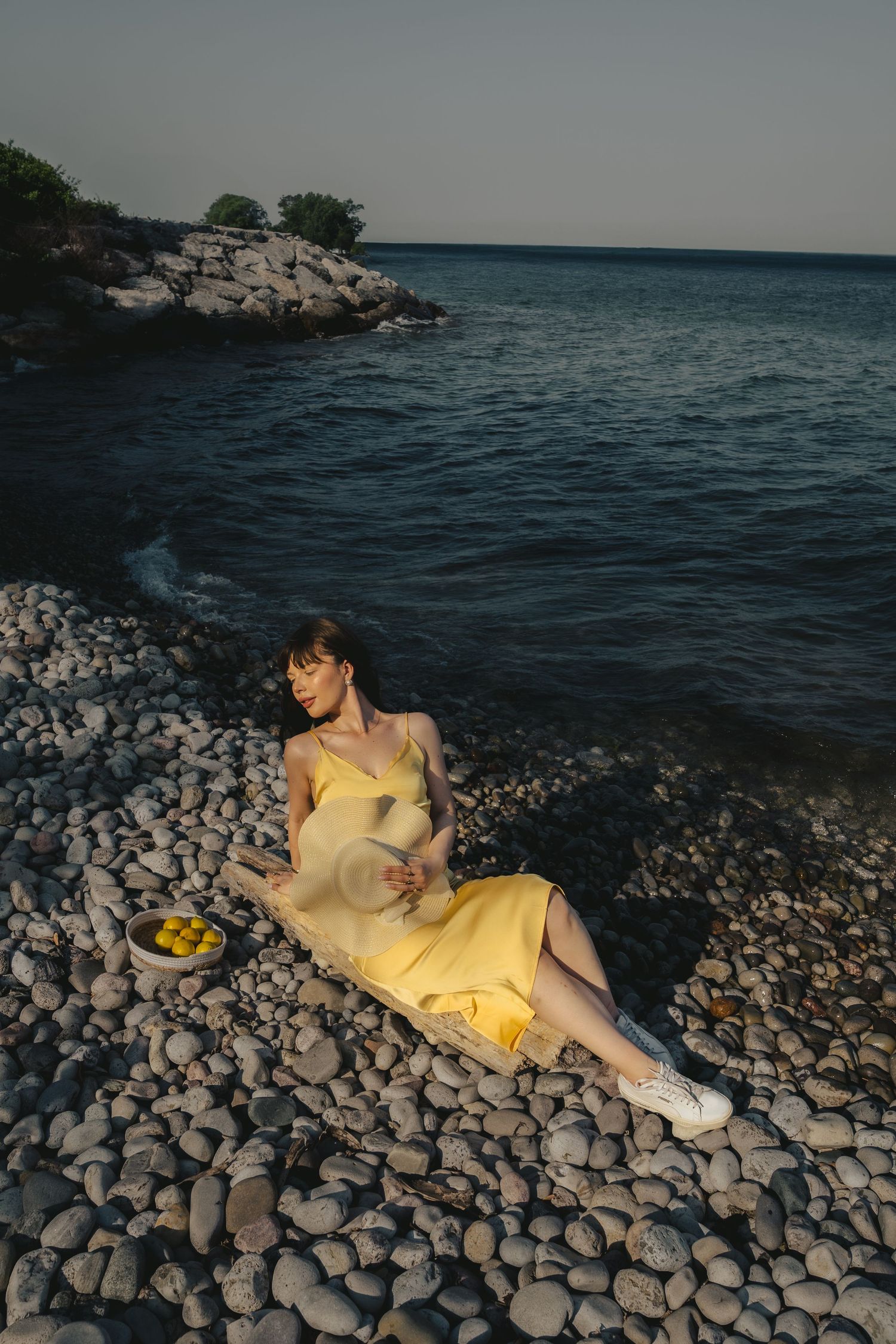 Person in yellow dress reclining on rocky shoreline at dusk.