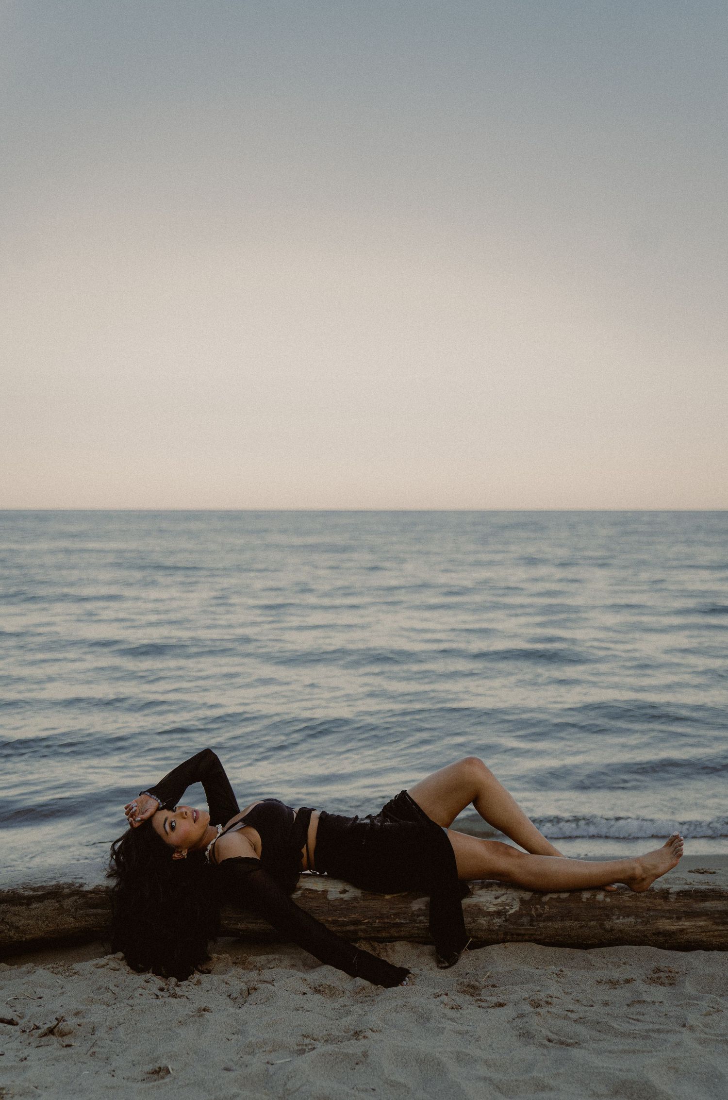 Figure in black clothing lying on beach sand with ocean horizon at dusk.