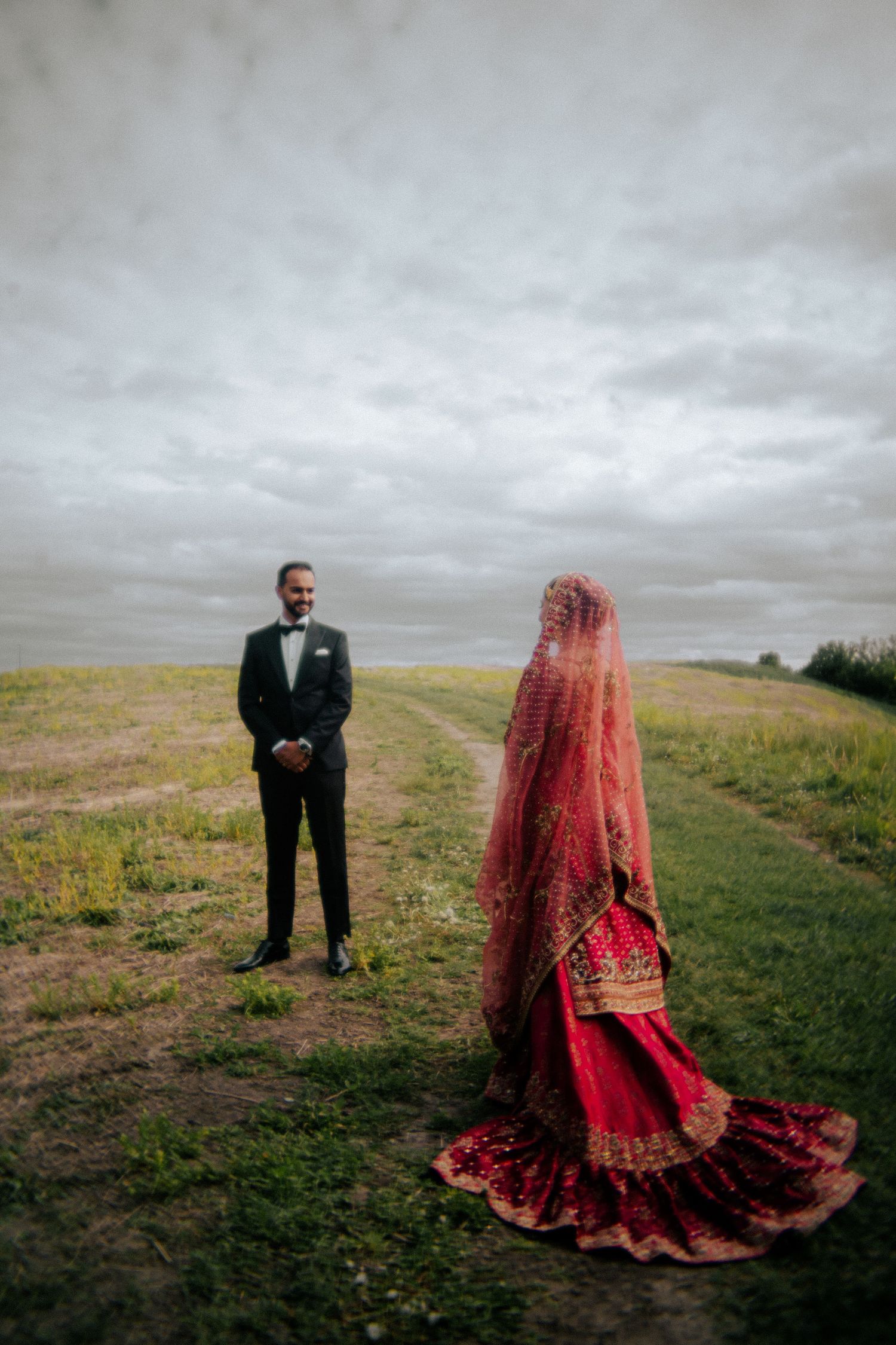 Wedding couple on hillside with traditional red dress against overcast sky.
