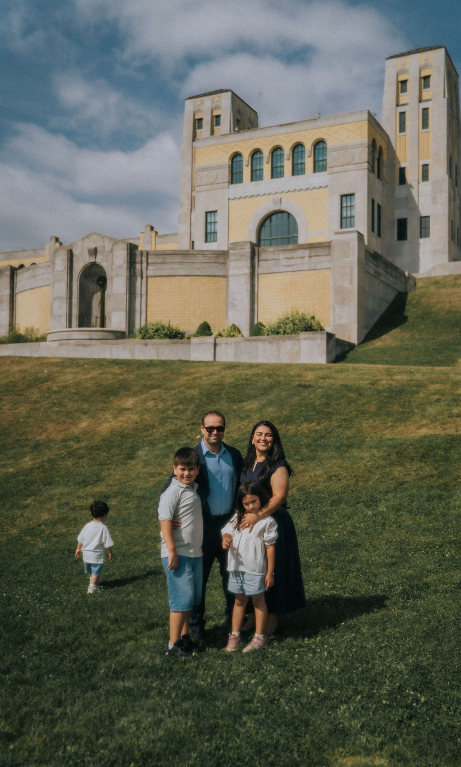 A family group stands on a grassy hillside in front of a grand architectural building with arched windows.