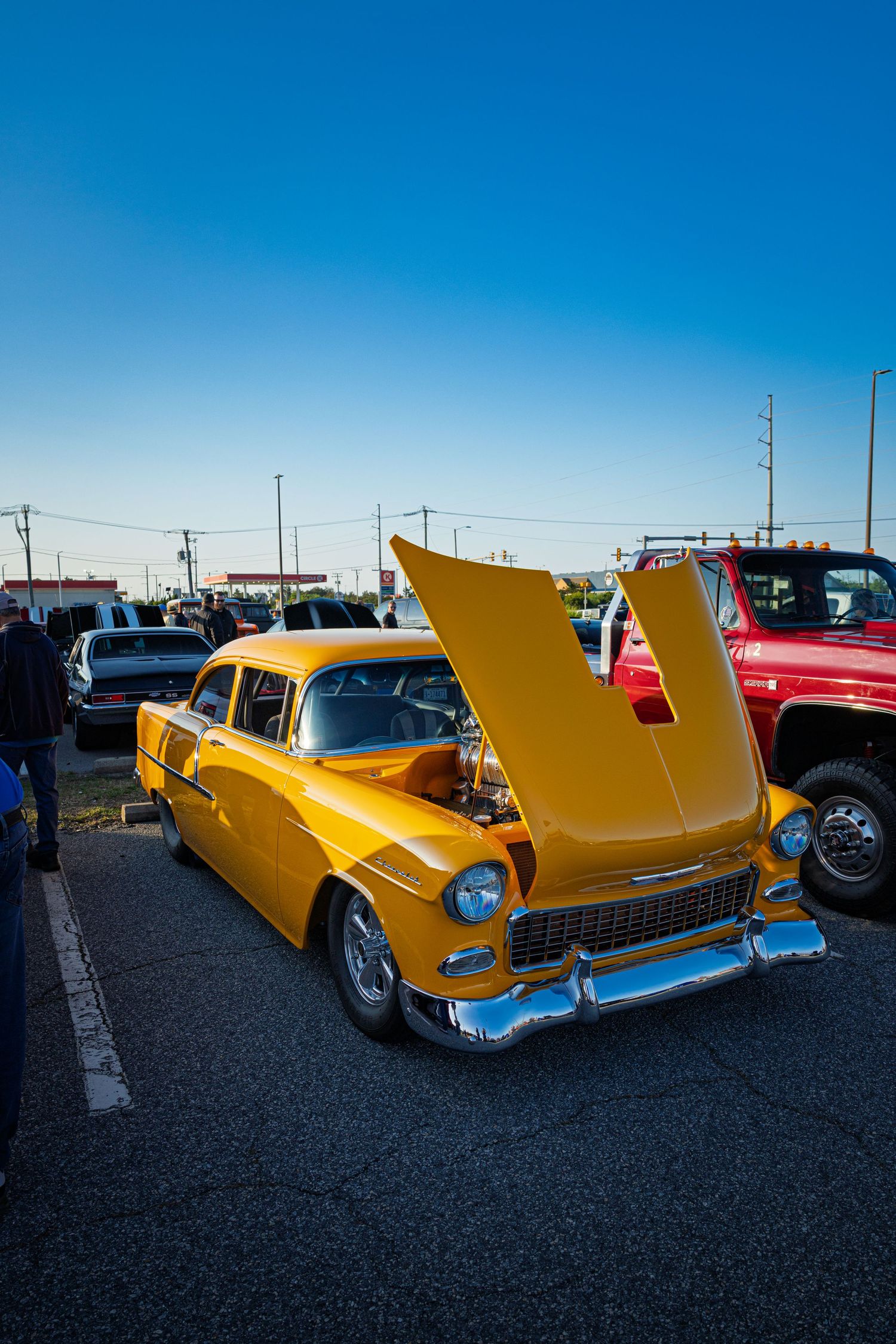 A classic yellow 1955 Chevy Bel Air with open hood on display at an outdoor car show under a clear blue sky.