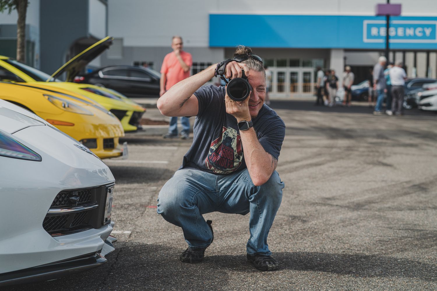 Photographer crouching to take photos at a car show with yellow and white vehicles in the background.