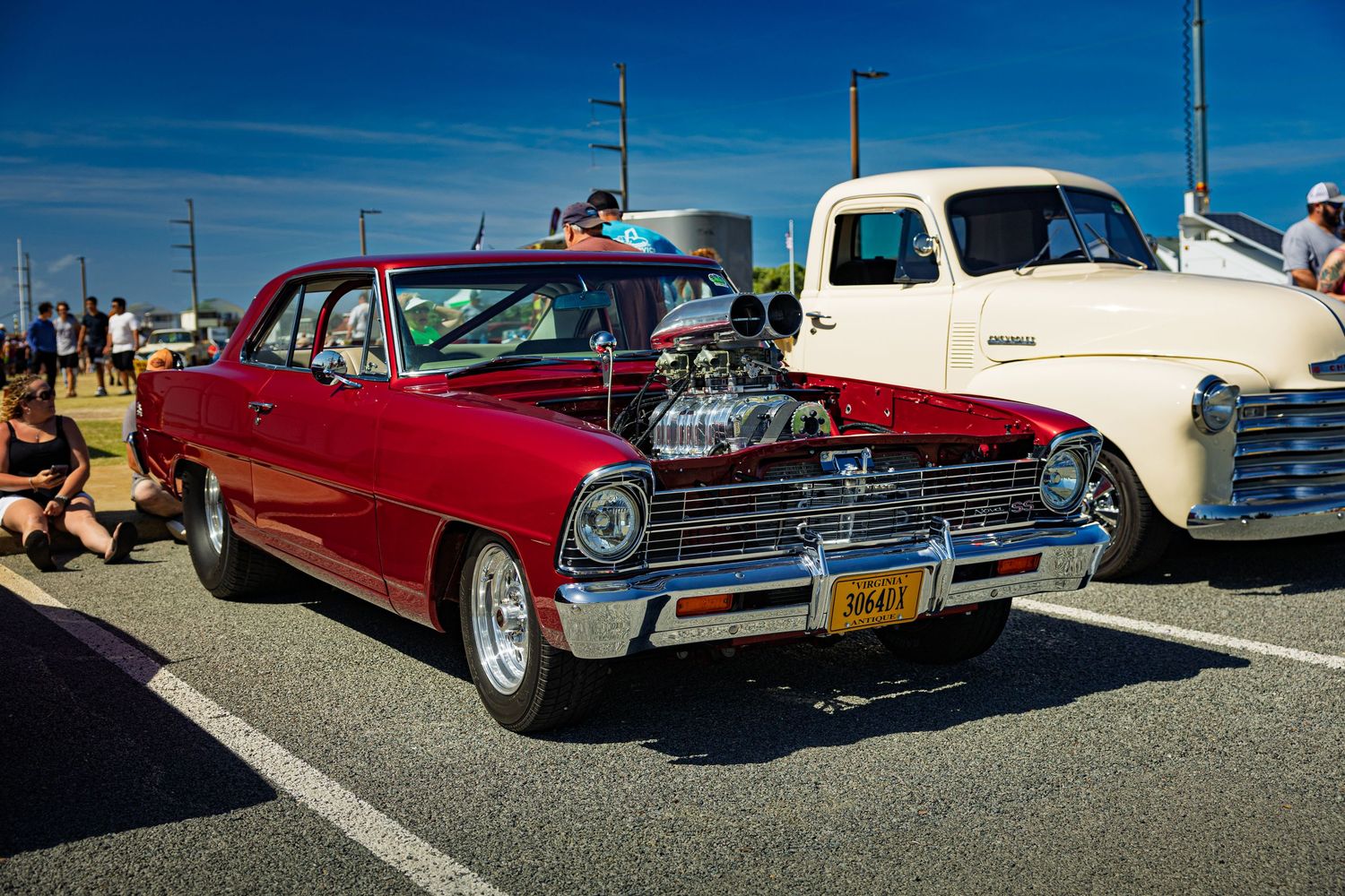 A classic red Chevrolet Nova with chrome trim and custom engine sits parked at a car show under bright blue skies.