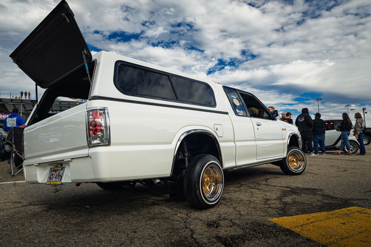 White lowrider pickup truck with gold wheels and hydraulics displayed at car show under cloudy sky.