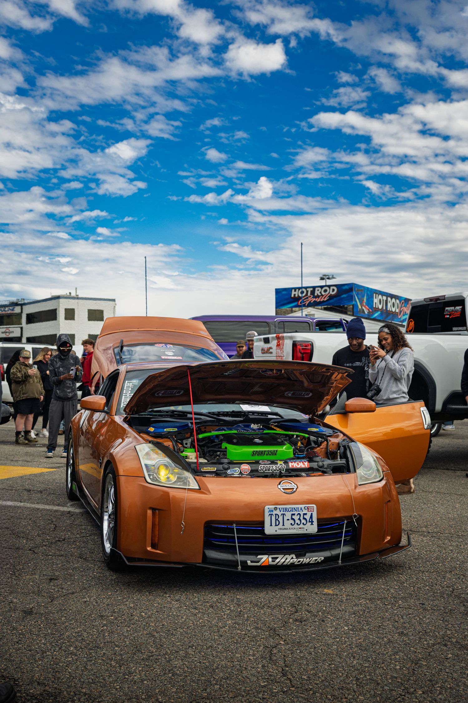 Custom orange lowrider car with chrome wheels displayed at an outdoor car show under blue skies with white clouds.