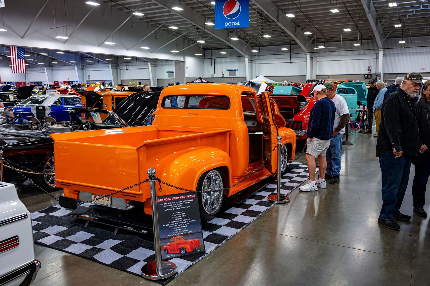 Custom orange pickup trucks on display at an indoor automotive show with black and white checkered flooring.