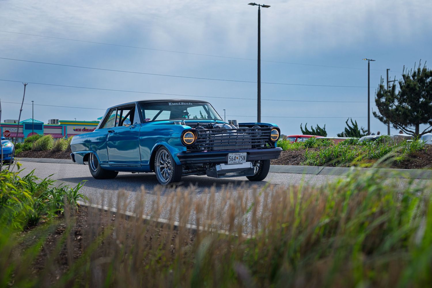 Classic blue vintage car parked on wet pavement with grass in foreground and cloudy sky in background.