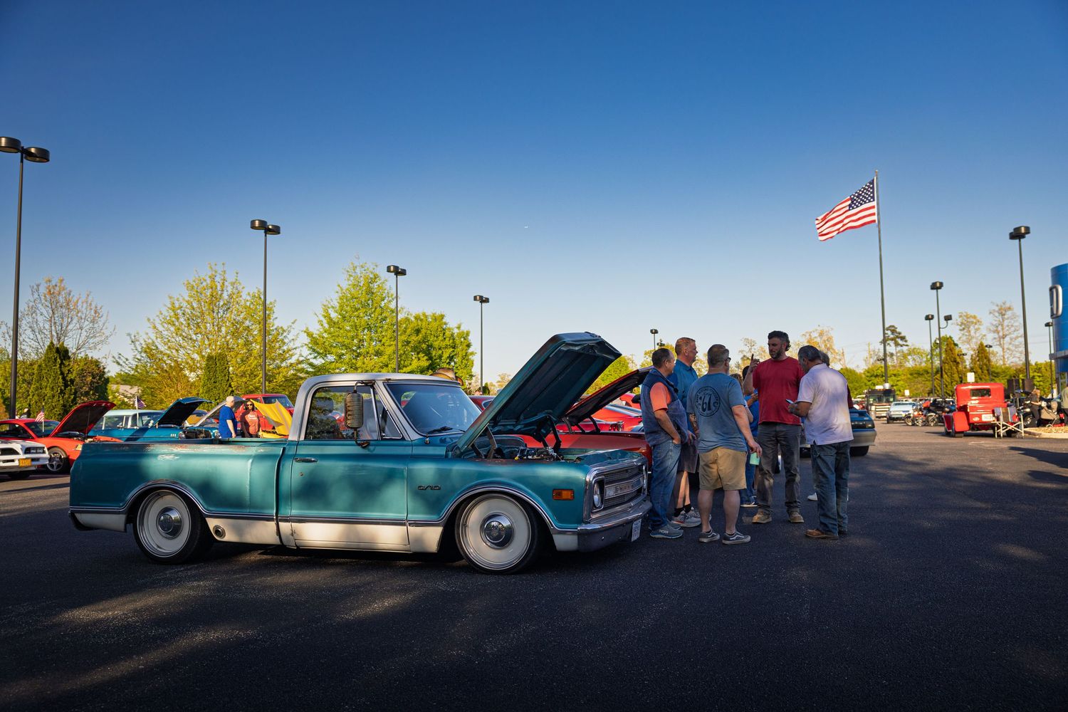 Group of people admiring a classic teal pickup truck with open hood at an outdoor car show under clear blue sky.
