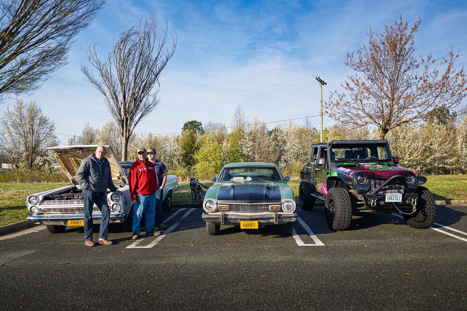 A panoramic view of parked cars and people gathered at an outdoor event on a sunny spring day.