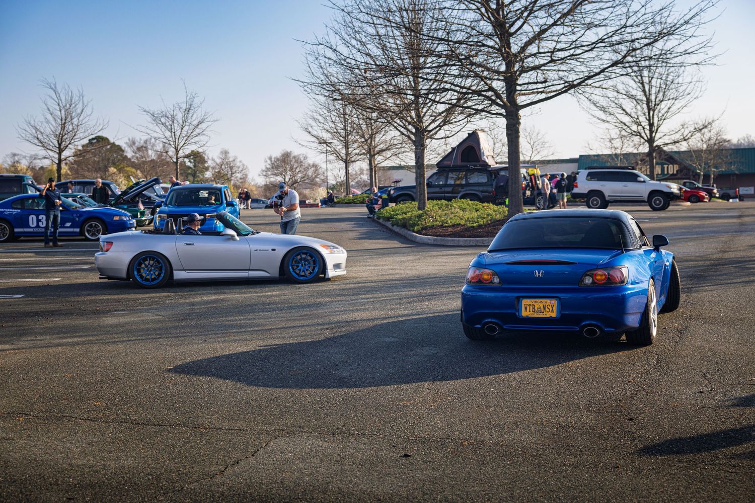 Two modified sports cars, one silver and one blue, parked in a lot with bare winter trees in the background.