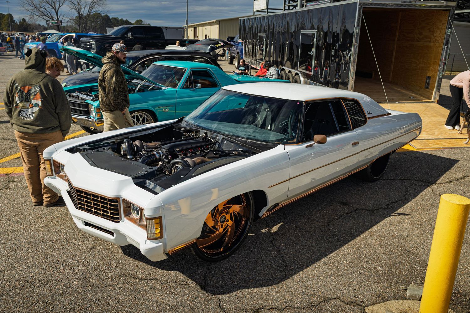 A customized white Chevrolt lowrider with bronze wheels displayed at a car show next to a turquoise classic car.