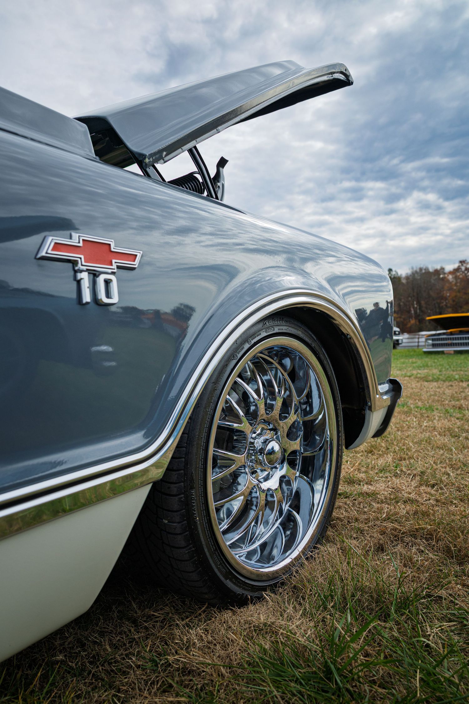 Close-up shot of a custom C10 truck's chrome wheels and silver body panels against a cloudy sky and grassy field.