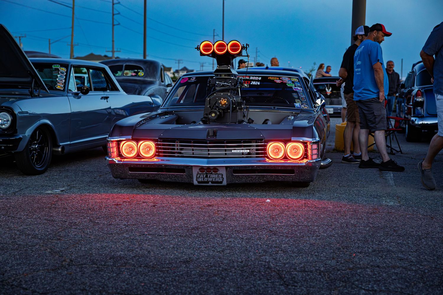 A row of classic cars with glowing red taillights parked at dusk during a car meet.
