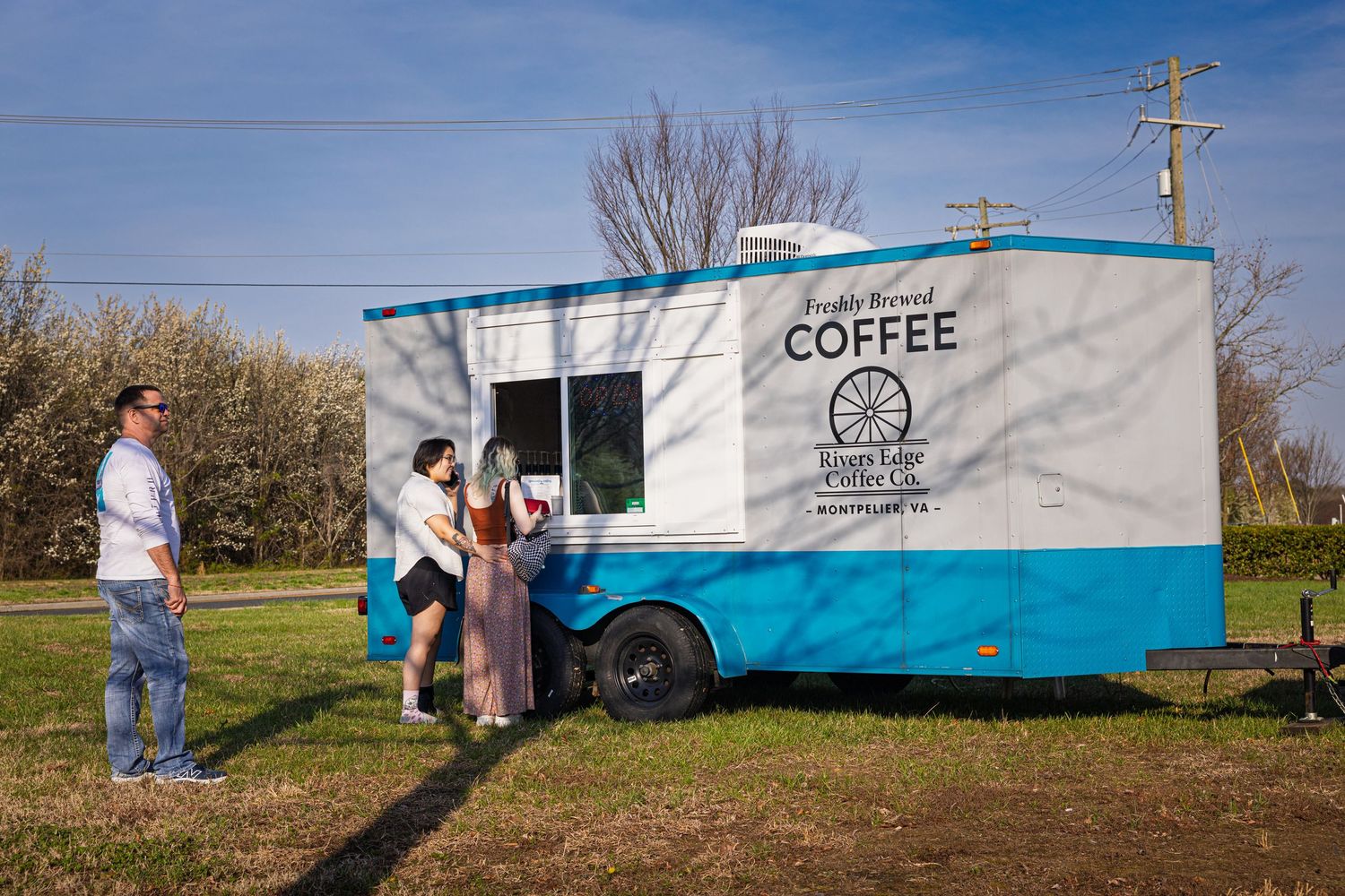 A mobile coffee trailer painted in turquoise and white serves customers in a sunny outdoor setting.