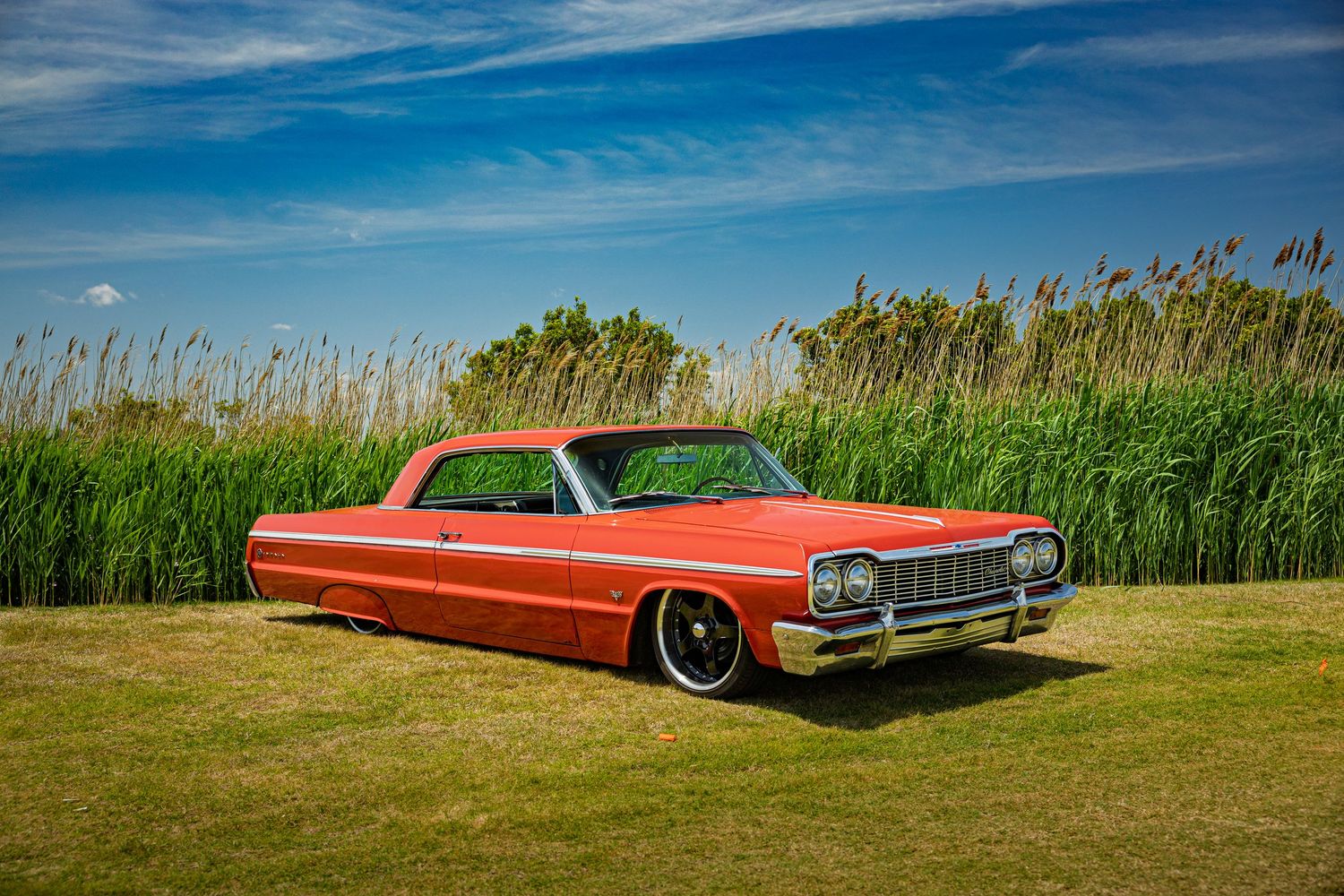 A bright red orange Chevrolet Impala lowrider sits in a grassy field against a vibrant blue sky.