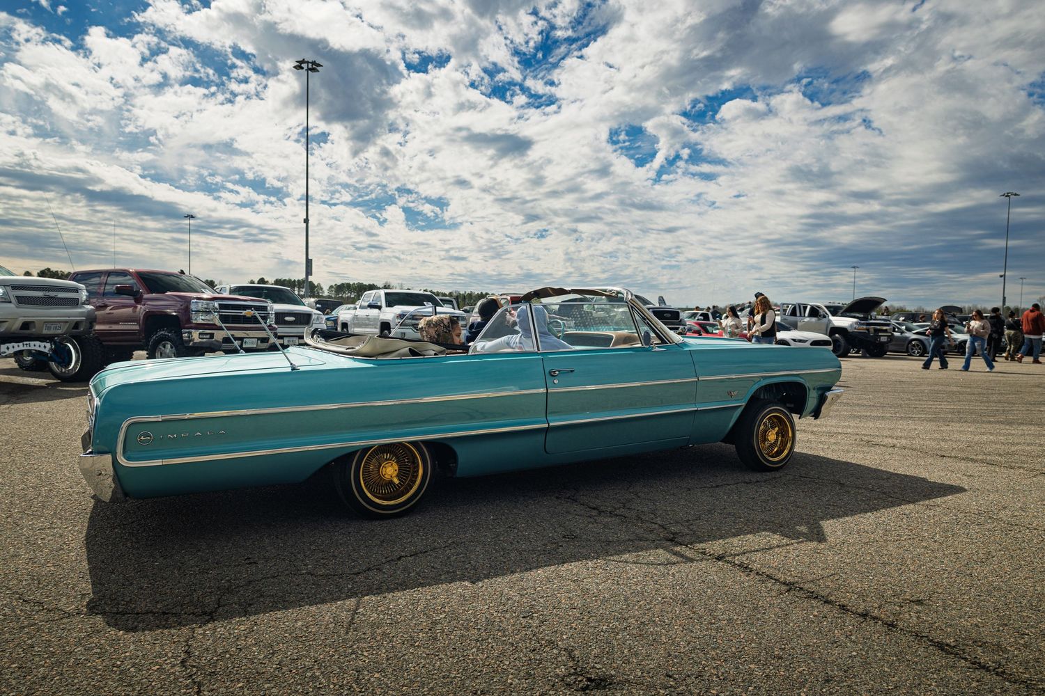 Row of vintage lowrider cars in turquoise and metallic colors showcased at a car meet under cloudy skies.