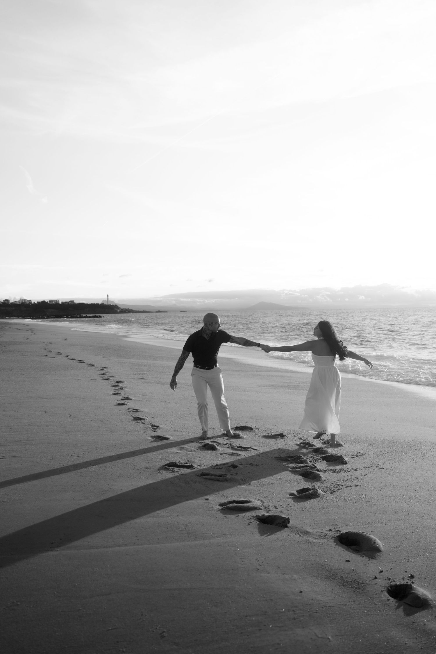 Black and white romantic couple holding hands walking along a beach shoreline with footprints in sand.