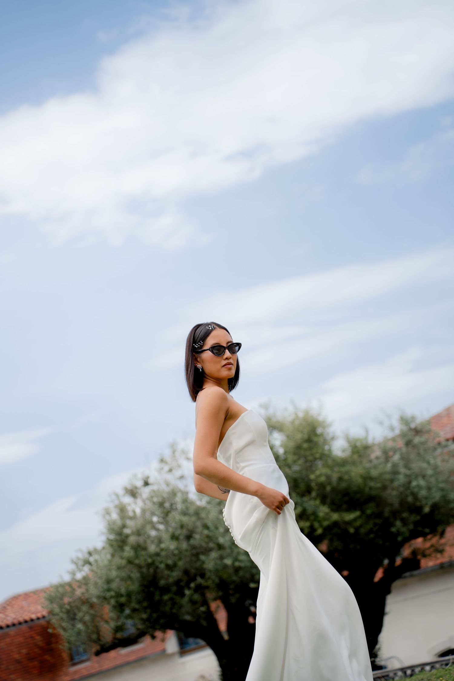Stylish bride in a white strapless gown and cat-eye sunglasses poses against a blue sky with olive trees.