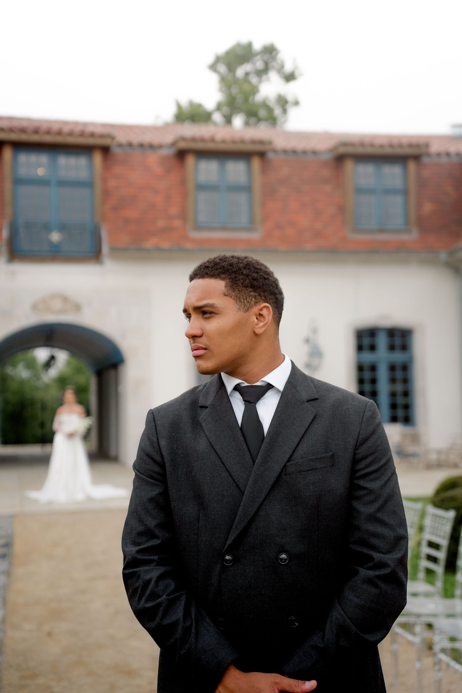 Groom in black double-breasted suit standing at outdoor wedding venue with bride visible in background archway.