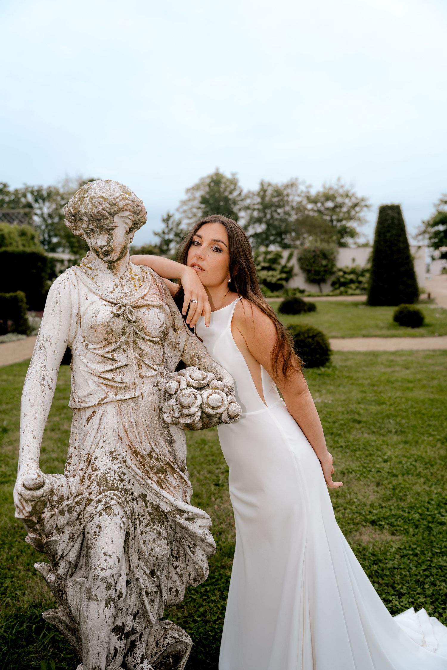 Elegant bride in fitted white dress leaning against antique stone goddess sculpture outdoors.