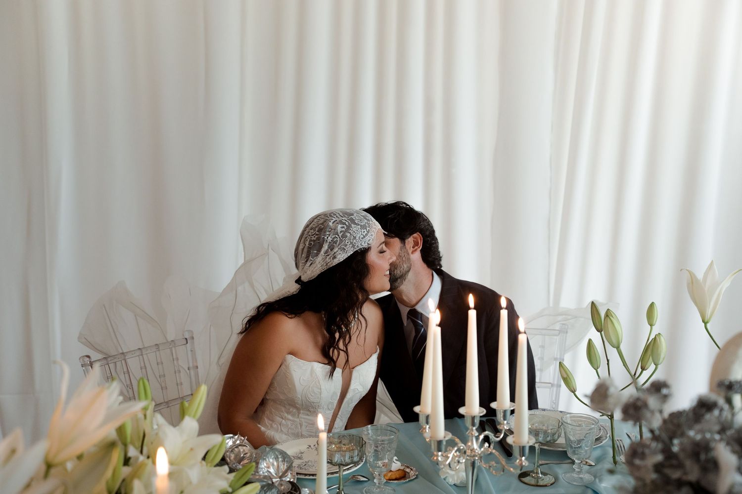 Bride and groom share a romantic kiss at a candlelit wedding reception table adorned with white lilies.
