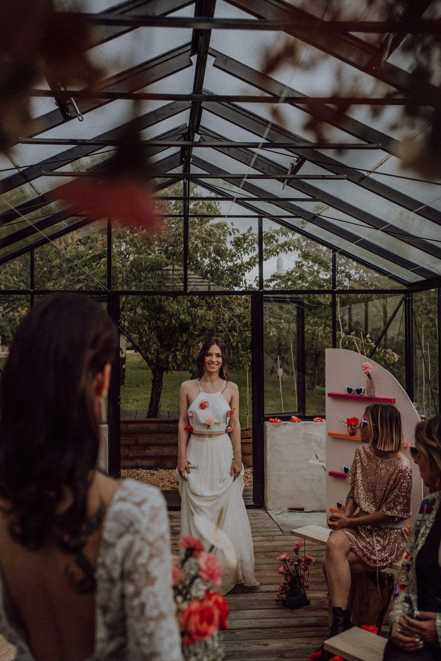 Bride in white dress walking toward her partner during an intimate greenhouse wedding ceremony with guests.
